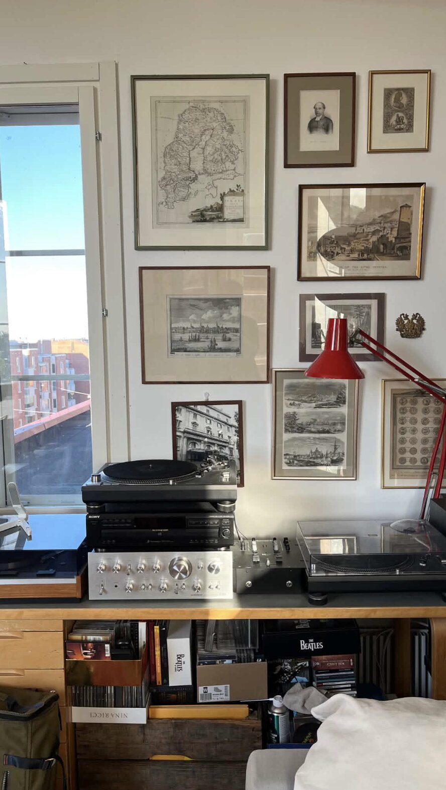 Mur de cartes anciennes et gravures encadrées, lampe rouge articulée, platine et ampli en dessous