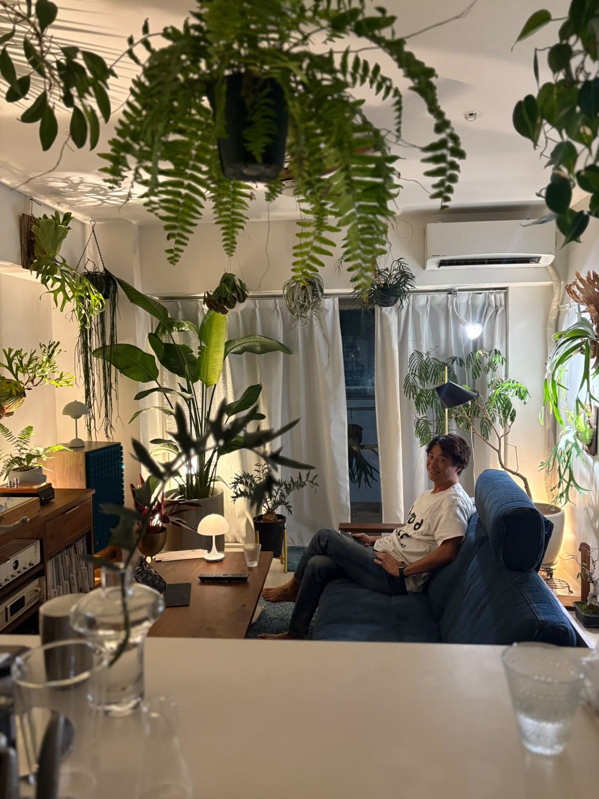 Leo assis sur le canapé en jean, souriant, entouré de plantes et de son mur de disques